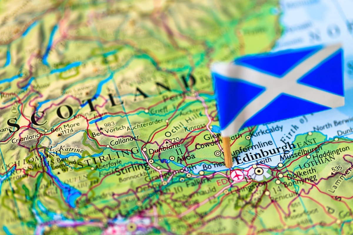 This image shows a close-up of a colourful, detailed topographical map focused on the area of Scotland, UK. Edinburgh is clearly marked, with a miniature Scottish flag placed on the city, indicating its location. Surrounding geographical features and cities such as Stirling and areas like Loch Lomond and The Trossachs National Park are also visible. The map's detail includes contour lines indicating terrain and elevation changes, roadways, and bodies of water, providing both geographical and political information.